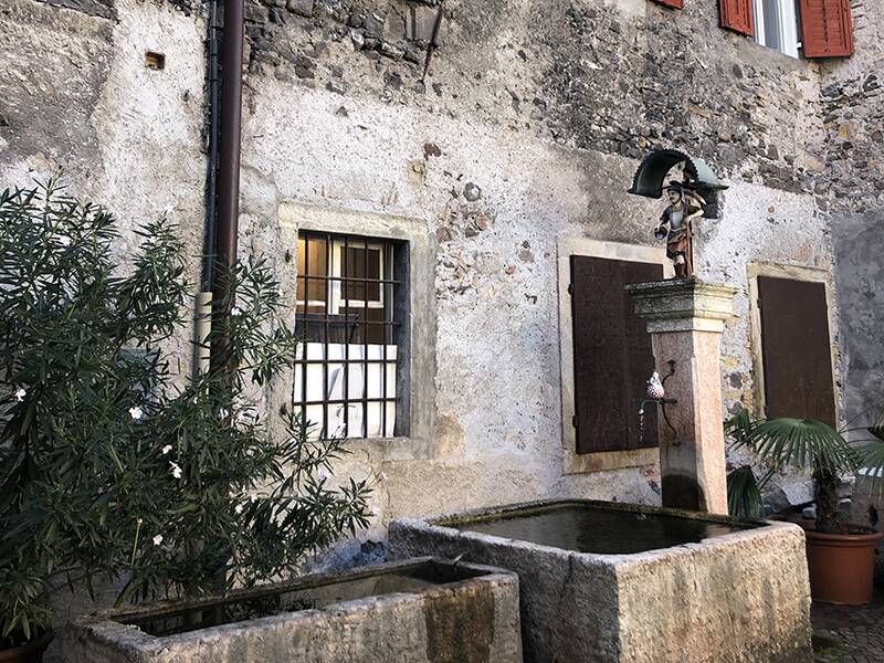 Ein steinerner Brunnen mit einer Skulptur auf einem Sockel steht vor einer alten, verputzten Steinwand mit mehreren Fenstern und einem Busch an der Seite.
