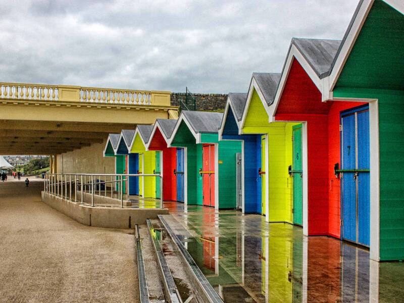 Bunte Strandhütten in verschiedenen Farben stehen entlang einer Promenade neben einer überdachten Struktur.