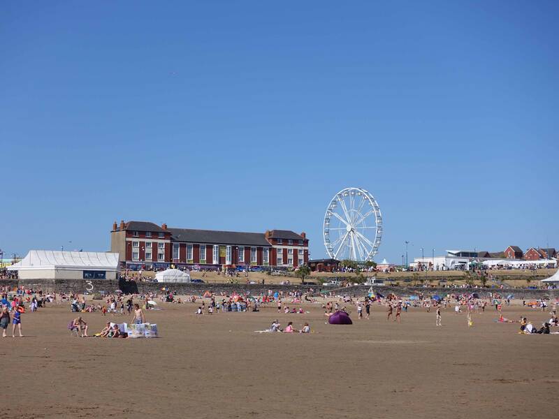 Zahlreiche Menschen genießen einen sonnigen Tag am Strand, während im Hintergrund ein Riesenrad und Gebäude zu sehen sind.