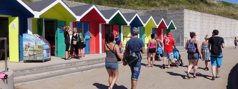 Mehrere Menschen spazieren auf einem sonnigen Weg an einer Reihe bunter Strandhütten vorbei.