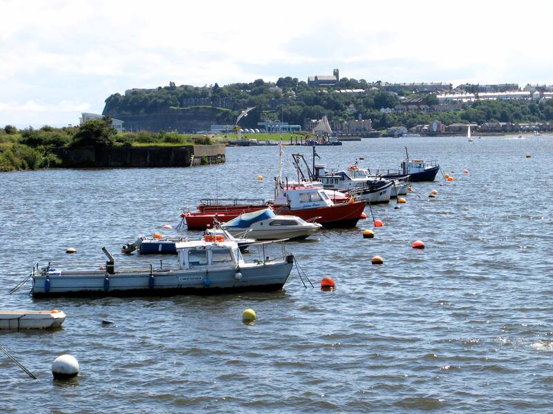 Mehrere Boote liegen im Wasser vertäut, wobei im Hintergrund eine hügelige Landschaft mit Gebäuden sichtbar ist.