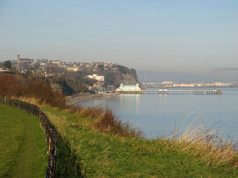 Eine Küstenlandschaft mit einem langen Pier und Häusern auf einem Hügel im Hintergrund, umgeben von ruhigem Wasser und einem grasbewachsenen Weg im Vordergrund.