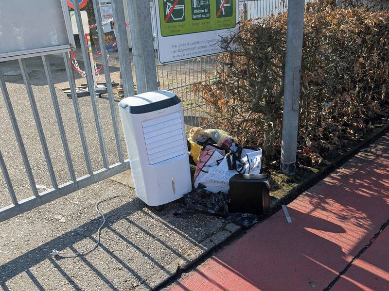 Neben einem metallenen Zaun steht ein Luftentfeuchter und mehrere Taschen und Müll sind daneben auf dem Boden abgestellt.