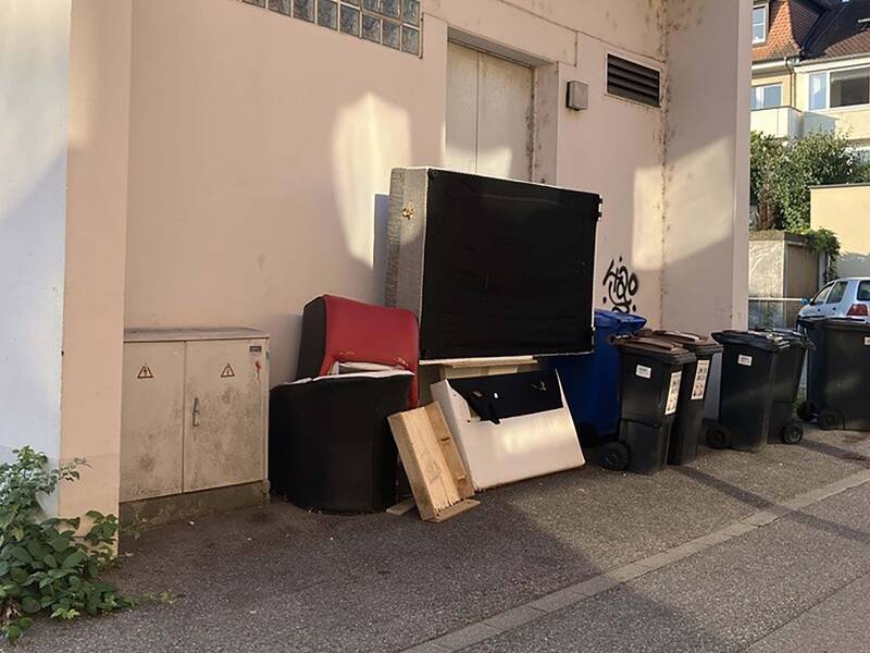 Vor einem Gebäude stehen mehrere Mülltonnen sowie sperrige Abfälle, darunter ein roter Sessel und Holzplatten.