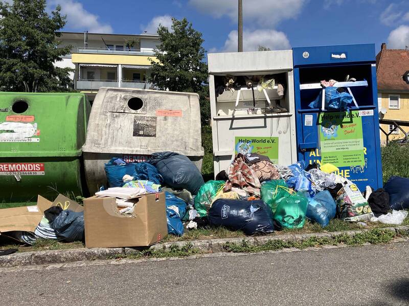 Vor mehreren Kleidercontainern und Altglascontainern liegen zahlreiche Müllsäcke und Kartons auf dem Boden.