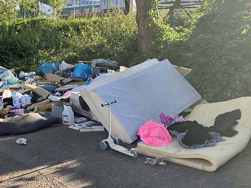 Am Rand einer Straße liegt eine große Menge an weggeworfenem Müll, einschließlich Möbelteilen, Kleidungsstücken und verschiedenen Gegenständen, in der Nähe von Büschen.