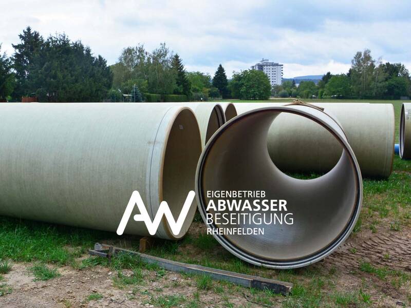Große graue Abwasserrohre liegen nebeneinander auf einer grasbewachsenen Fläche mit Bäumen und einem Gebäude im Hintergrund; im Vordergrund ist das Logo der Eigenbetrieb Abwasserbeseitigung Rheinfelden zu sehen.