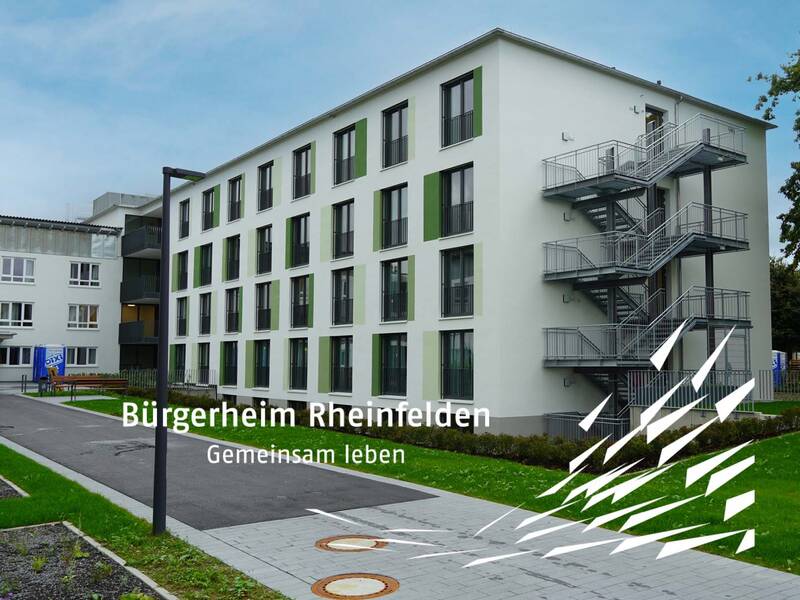 Das Bild zeigt ein modernes, mehrstöckiges Gebäude mit einem außenliegenden Treppenaufgang und der Aufschrift "Bürgerheim Rheinfelden - Gemeinsam leben" im Vordergrund.