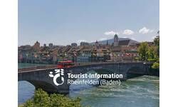 Eine Brücke mit wehenden Fahnen überspannt einen Fluss, im Hintergrund sind historische Gebäude und ein Hügel zu sehen, mit der Aufschrift "Tourist-Information Rheinfelden (Baden)".