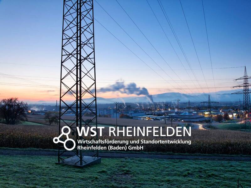 Das Bild zeigt eine industrielle Landschaft bei Sonnenuntergang mit Strommasten im Vordergrund und einem Schriftzug "WST Rheinfelden" darüber.