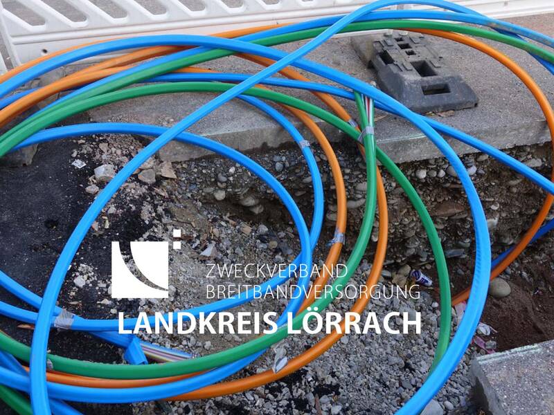 Bunte Kabel liegen auf einem aufgerissenen Boden mit Kies und Erde, über dem das Logo und der Schriftzug des Zweckverbands Breitbandversorgung Landkreis Lörrach eingeblendet sind.