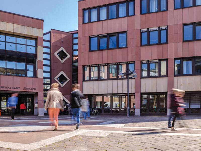 Mehrere Menschen gehen auf einem Platz vor dem Gebäude des Landratsamts Lörrach entlang.