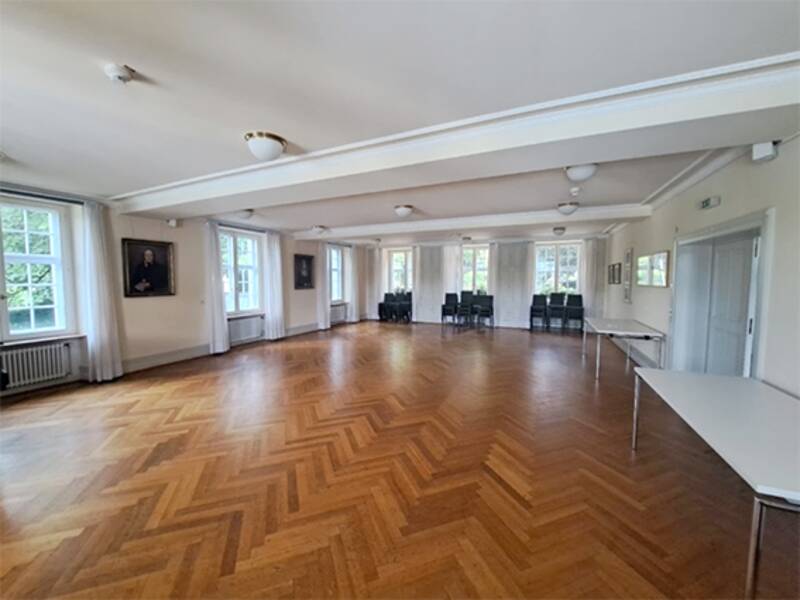 Ein leerer Raum mit Holzboden und großen Fenstern, mehreren Stühlen an einer Wand und einem großen Tisch an der Seite.