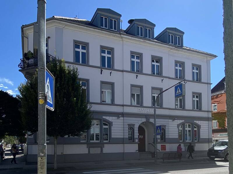 Das Gebäude des alten Rathauses mit 3 Stockwerken und vielen Fenstern, im Vordergrund die Friedrichstraße mit Verkehrszeichen und Bäumen