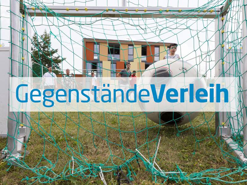 Ein großer aufblasbarer Ball liegt in einem Fußballtor auf einer Wiese, im Hintergrund sind Menschen und ein modernes, buntes Gebäude zu sehen, die über den Schriftzug "Gegenstände Verleih" schauen.