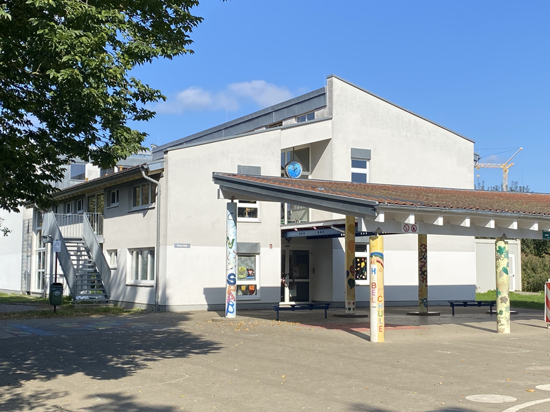 Ein modernes Gebäude mit weißer Fassade und einem überdachten Eingangsbereich, unterstützt von bunt bemalten Säulen, steht unter klarem, blauem Himmel.