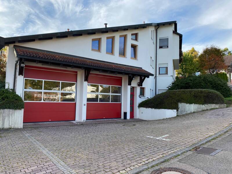 Ein modernes Gebäude mit roten Garagentoren und einem geneigten Pflasterweg davor, eingebettet in eine grüne, bewaldete Umgebung.