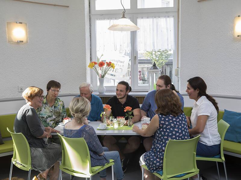 Eine Gruppe von sieben Personen sitzt um einen Tisch in einem gemütlichen Café und unterhält sich, während auf dem Tisch Getränke und Blumen stehen.