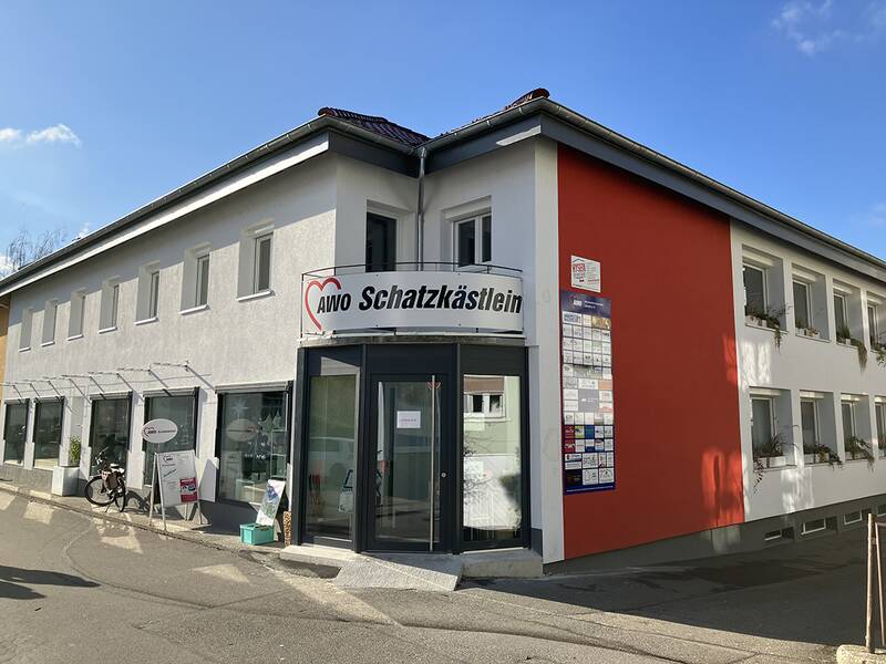 Das Bild zeigt ein zweistöckiges Gebäude mit der Aufschrift "AWO Schatzkästlein" über dem Eingang, das graue und rote Fassaden hat und an einer sonnigen Straße liegt.