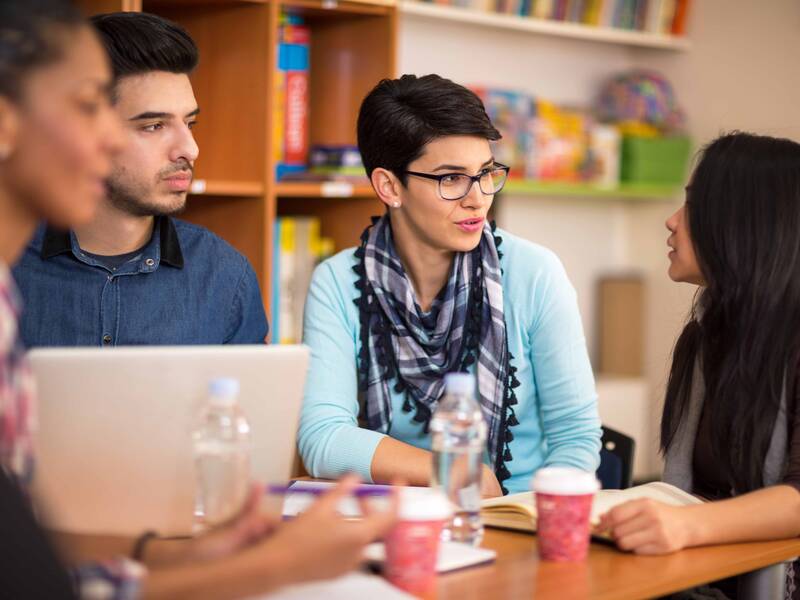 Vier Personen sitzen um einen Tisch und führen ein Gespräch, während im Hintergrund Bücherregale sichtbar sind.