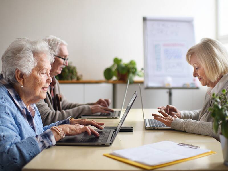 Drei ältere Menschen sitzen an einem Tisch und arbeiten an Laptops in einem hellen Raum mit Pflanzen.