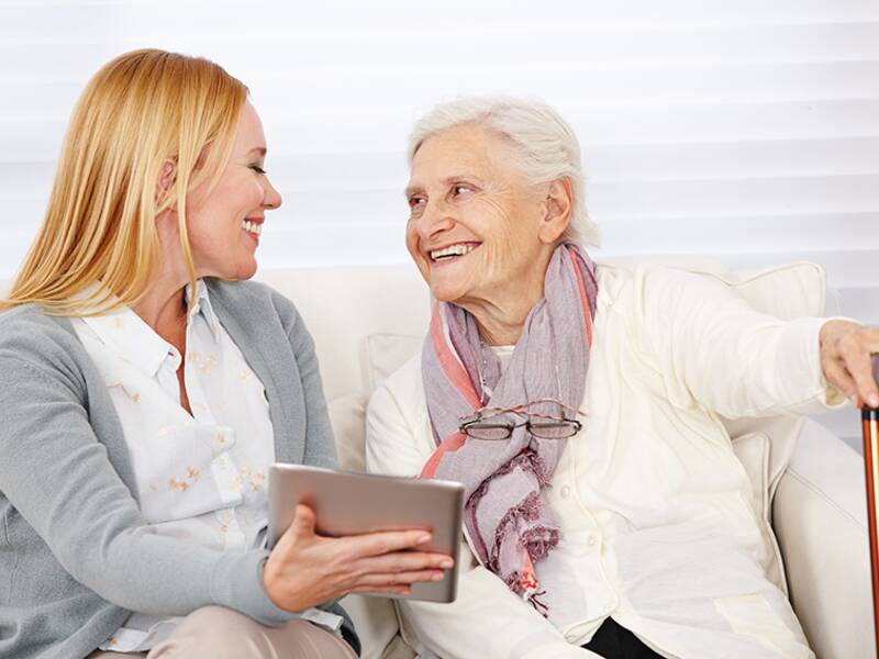 Eine jüngere Frau und eine ältere Frau sitzen lächelnd auf einem Sofa, während die jüngere Frau ein Tablet hält.