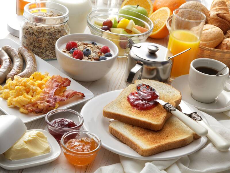 Ein reichhaltiges Frühstück mit Toast, Marmelade, Speck, Rührei, Würstchen, frischem Obst, Müsli, Saft und Kaffee ist auf einem Tisch angerichtet.