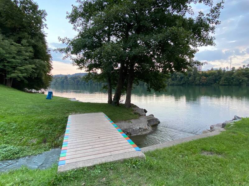 Ein Holzsteg führt über eine Bach, links und rechts Liegewiesen am Rhein und große Laubbäume