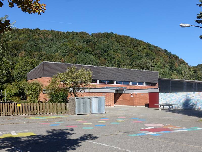 Vor einem dicht bewachsenen Hügel steht ein Backsteingebäude mit flachem, dunklem Dach, umgeben von einem breiten Vorplatz mit bunten Markierungen und einem leeren Parkplatz.
