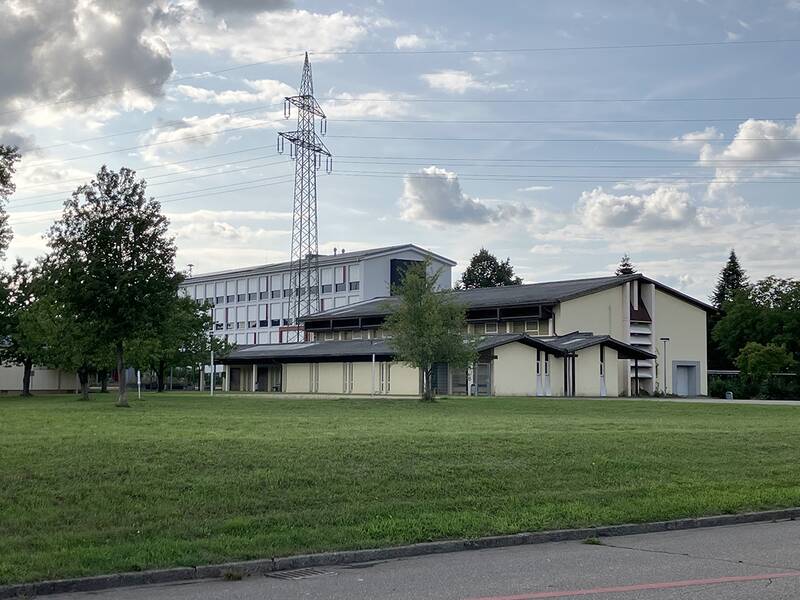 Ein modernes, weiß-gelbes Gebäude steht auf einer großen grünen Wiese mit einem Strommast und Bäumen im Hintergrund unter einem bewölkten Himmel.