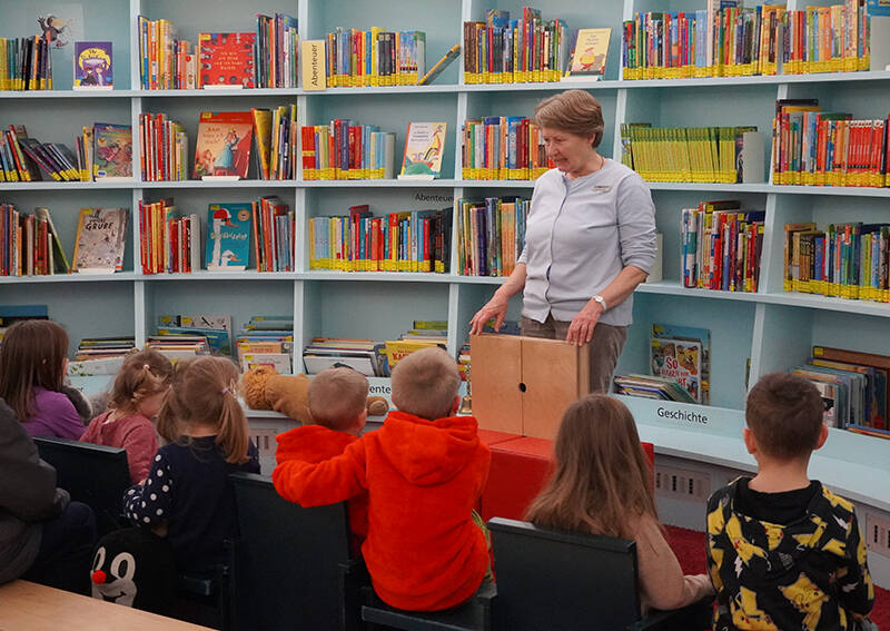 Eine Frau liest einer Gruppe von Kindern in einer Bibliothek mit vielen bunten Büchern im Hintergrund vor.