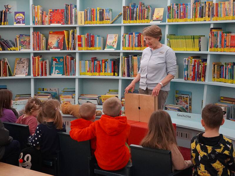 Eine Frau liest einer Gruppe von Kindern in einer Bibliothek mit vielen bunten Büchern im Hintergrund vor.