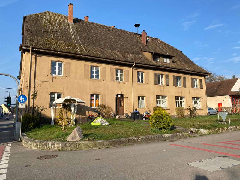 Ein großes, zweistöckiges Haus mit mehreren Fenstern und einem verwitterten Dach steht an einer Straßenecke, umgeben von kleinen Pflanzen und einem Weg, der zu einer Kreuzung führt.