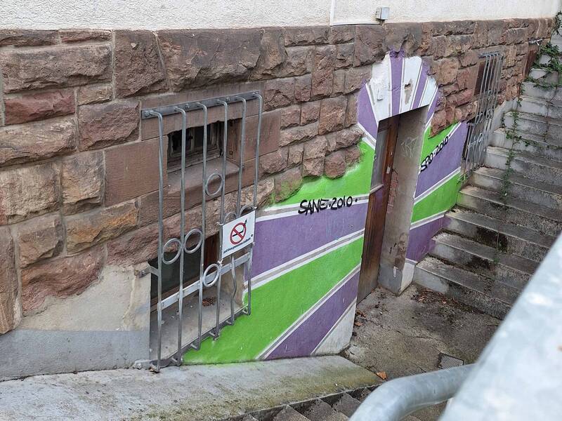 Die Treppe führt zur Tür des Jugendkellers, ein vergittertes Fenster und ein Graffiti an der Hauswand