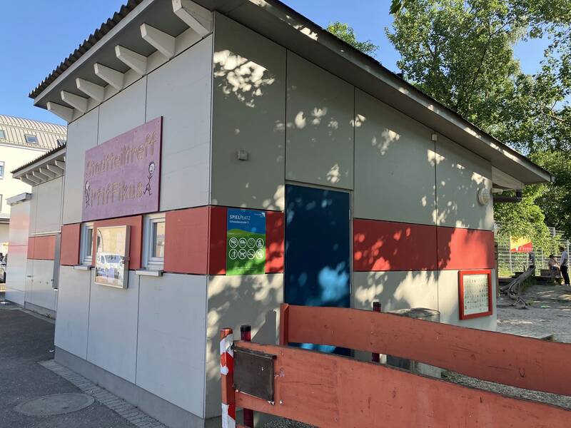 Gebäude des Stadtteiltreffs Pfiffikus: Graues, niedriges Häuschen, dass mit einem roten, es umringenden Streifen geschmückt ist. 