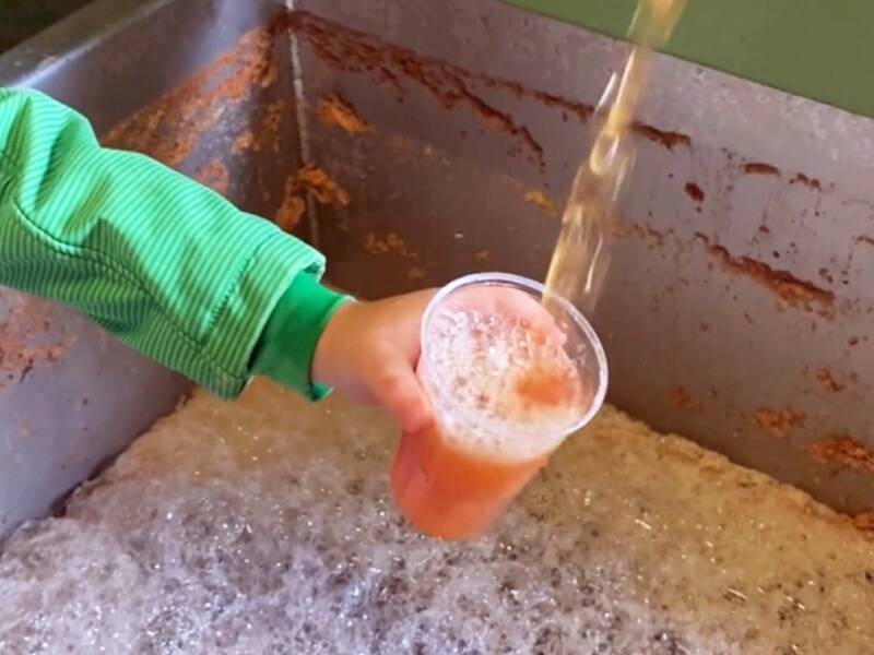 Eine Person mit grüner Jacke hält einen Becher, der mit einer schaumigen, orangefarbenen Flüssigkeit gefüllt wird, über einem Behälter voller Schaum.