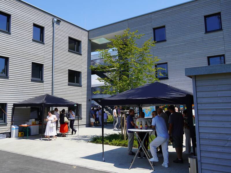 Auf dem Bild sind Menschen versammelt und plaudern unter Pavillons vor einem modernen, mehrstöckigen Gebäude an einem sonnigen Tag.