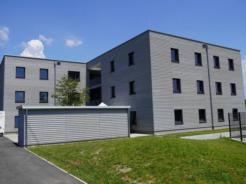 Das Bild zeigt ein modernes, mehrstöckiges Gebäude mit grauer Fassade, umgeben von einer Grünfläche und blauem Himmel im Hintergrund.