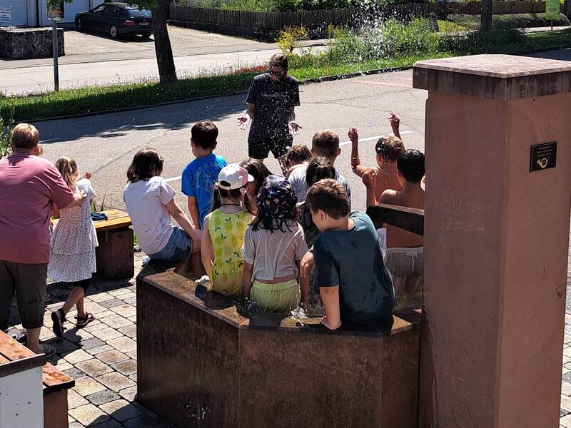 Eine Gruppe von Kindern sitzt und steht auf einem Brunnenrand und spielt mit Wasser, während ein Erwachsener in der Nähe steht und nass wird.