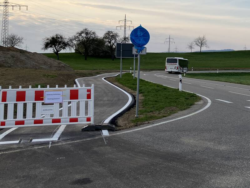 Ein neu gebauter Fahrradweg endet abrupt mit einer Absperrung und führt direkt auf eine stark befahrene Straße neben einem vorbeifahrenden Bus.
