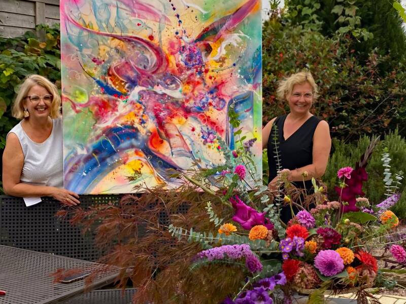 Petra Heck und Rita Bruckner mit einem großflächigen Gemälde und einem Blumenarrangement.