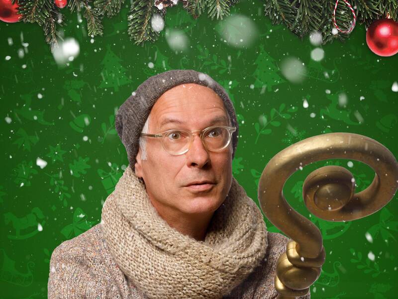 Ein Mann mit Brille, grauer Mütze und Schal hält einen goldenen Stab vor einem grünen Hintergrund mit Schneeflocken und Weihnachtsdekorationen.