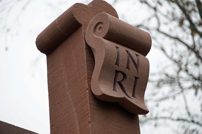 Ein Sandsteinkreuz mit einer Schriftrolle, auf der "INRI" eingraviert ist, vor einem Hintergrund aus blassgrauem Himmel und kahlen Baumästen.