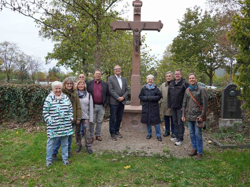 Eine Gruppe von Menschen steht lächelnd vor einem steinernen Kruzifix in einer grünen, parkähnlichen Umgebung.