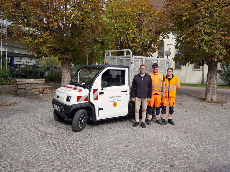 Drei Personen stehen in Arbeitskleidung neben einem kleinen weißen Nutzfahrzeug auf einem gepflasterten Platz mit herbstlichen Bäumen im Hintergrund.