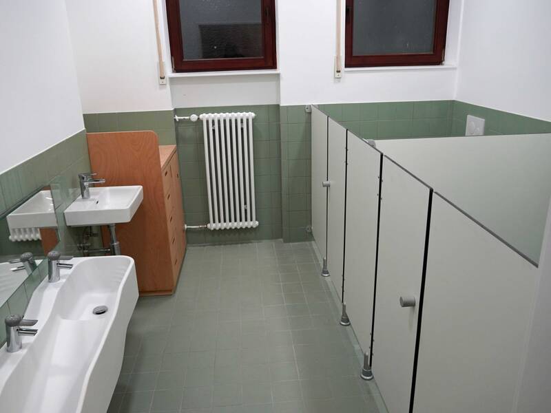 Ein schmaler Waschraum mit grünen Fliesen zeigt zwei Waschbecken mit einem Spiegel darüber, Holzmöbel, eine Toilette hinter Trennwänden und zwei Fenster.