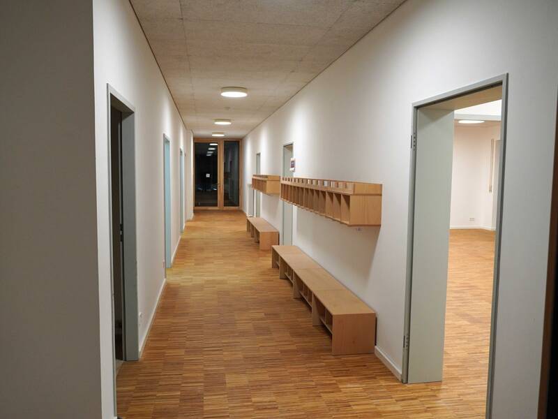 Ein langer, gut beleuchteter Flur mit Holzböden, offenen Türen zu beiden Seiten sowie Holzbänken und Fächern an der Wand.