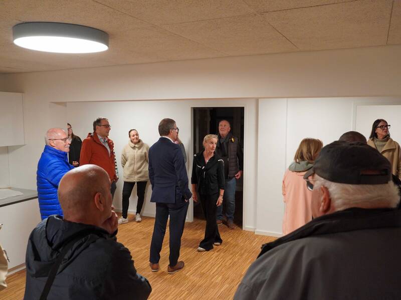 Eine Gruppe von Menschen steht in einem schlicht eingerichteten Raum mit Holzboden und unterhält sich.