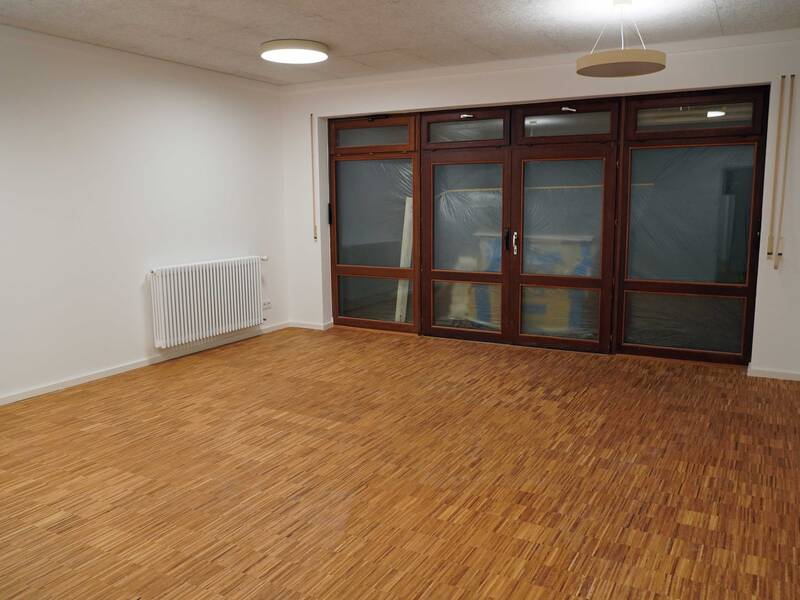 Ein leerer Raum mit Holzboden, weißen Wänden, einem Heizkörper an der linken Wand und großen, teilweise abgedeckten Fenstern auf der rechten Seite.