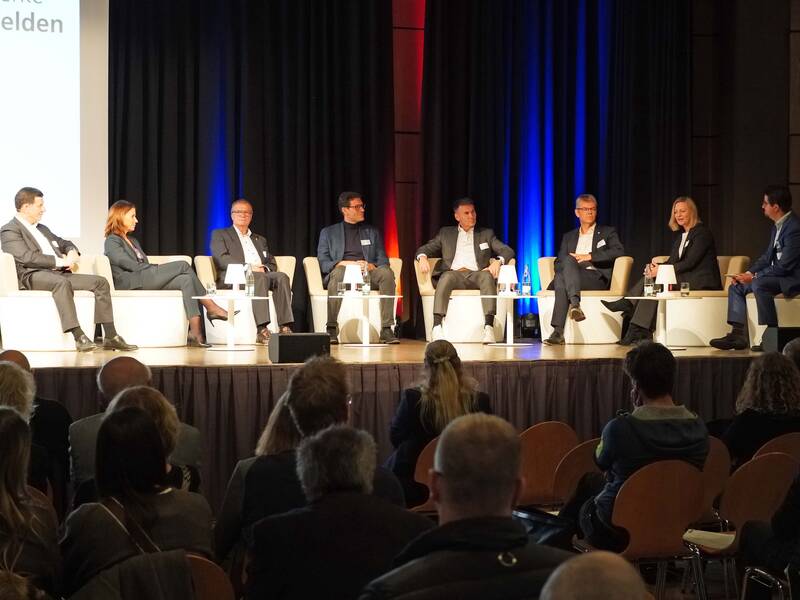 Eine Gruppe von sieben Personen sitzt in einer Podiumsdiskussion auf einer Bühne vor einem Publikum.